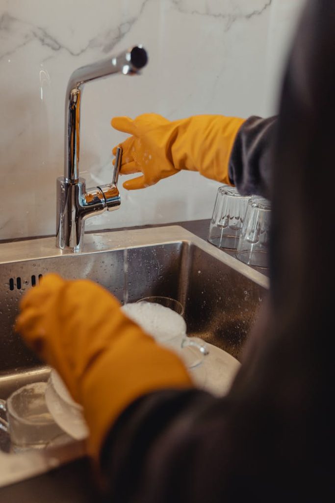 person-in-yellow-jacket-washing-hands-6195202 Close-up of a person washing dishes with yellow gloves in a kitchen sink.